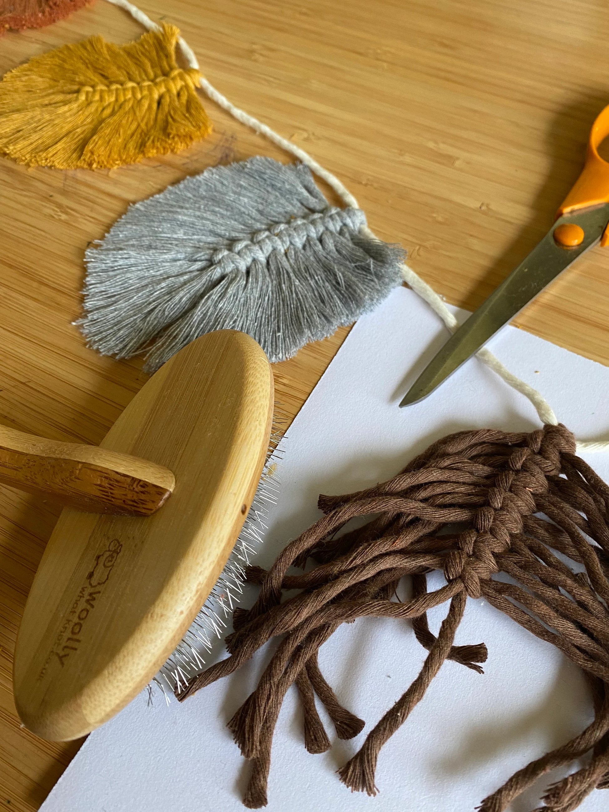 A crafting scene on a wooden surface features a Macra-Made-With-Love 100% cotton Autumn leaf garland - wall decor 180cm with macrame feathers in yellow, gray, and brown attached to a string. Nearby are a pair of orange scissors, a wooden brush tool, and a white sheet of paper.