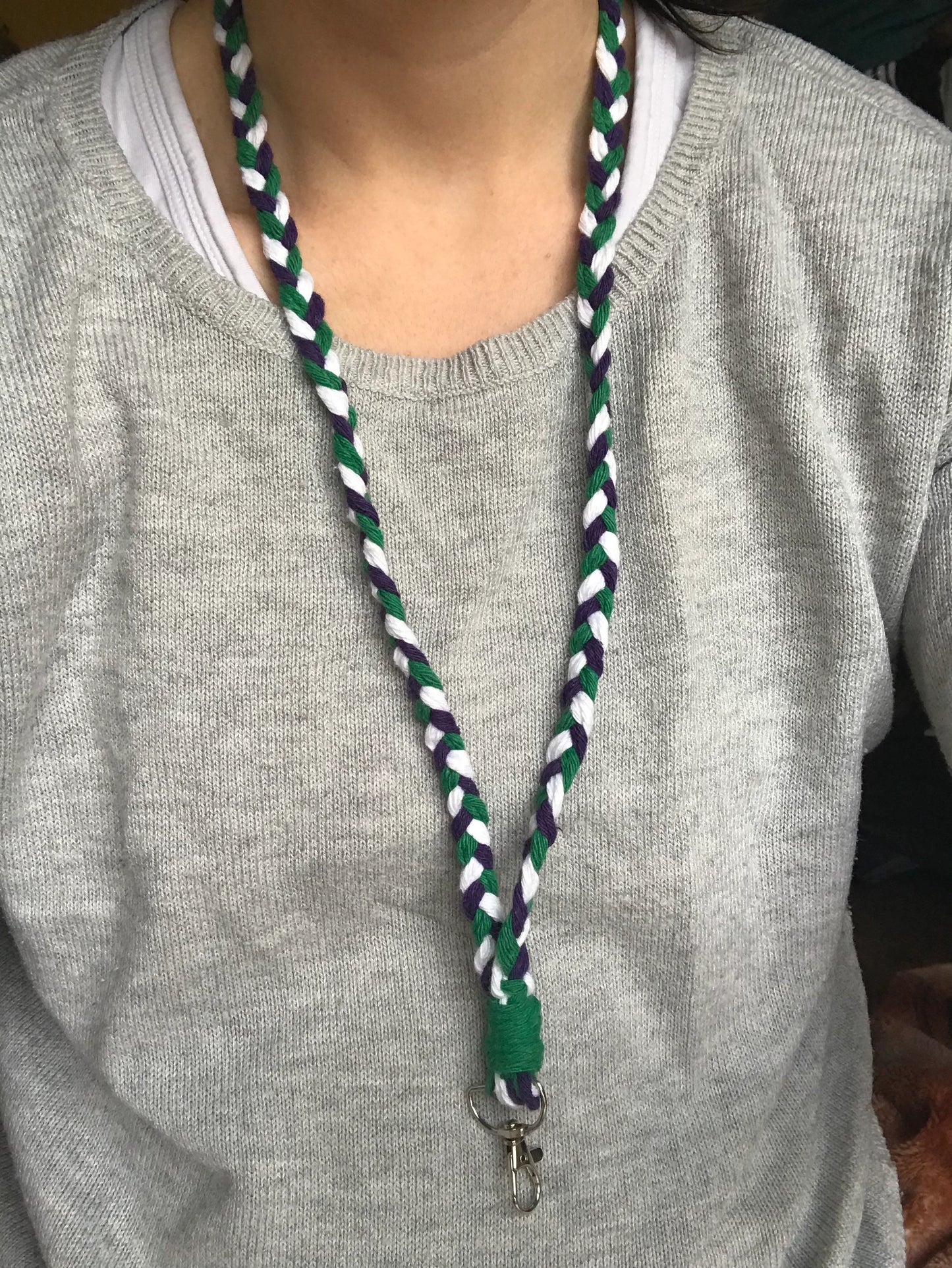 A person wearing a light gray sweater is shown from the shoulders down, showcasing an Eco friendly lanyard badge holder - 34” around their neck. The handmade lanyard by Macra-Made-With-Love is made of intertwined green, purple, and white strands, complete with a metal clip for an ID badge holder at the end.