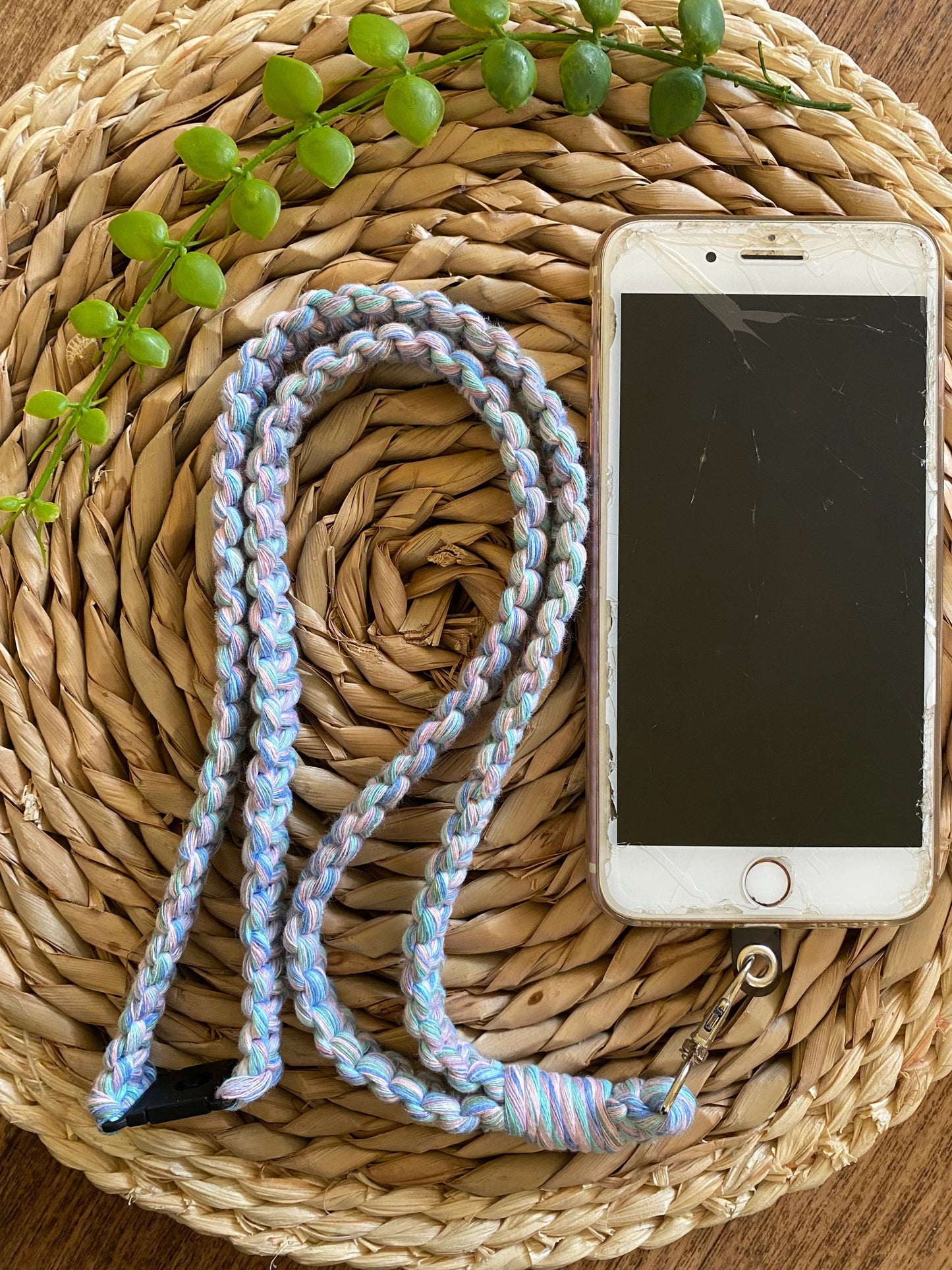 A cracked smartphone with a home button lies screen-up on a round straw mat. Attached to the phone is a Colourful knotted phone strap lanyard - 34” by Macra-Made-With-Love, made from recycled cotton cord, looped into a figure-eight shape. Small green succulent leaves rest on the mat near the phone and strap.