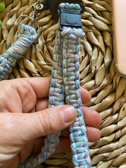 A hand is holding a Colourful knotted phone strap lanyard - 34” from Macra-Made-With-Love, made from a knitted, recycled cotton cord with a metal clip at the end. The lanyard is crafted from multicolored yarn in shades of blue, green, and purple. The background features a woven basket texture with part of a pink phone case visible on the right side.