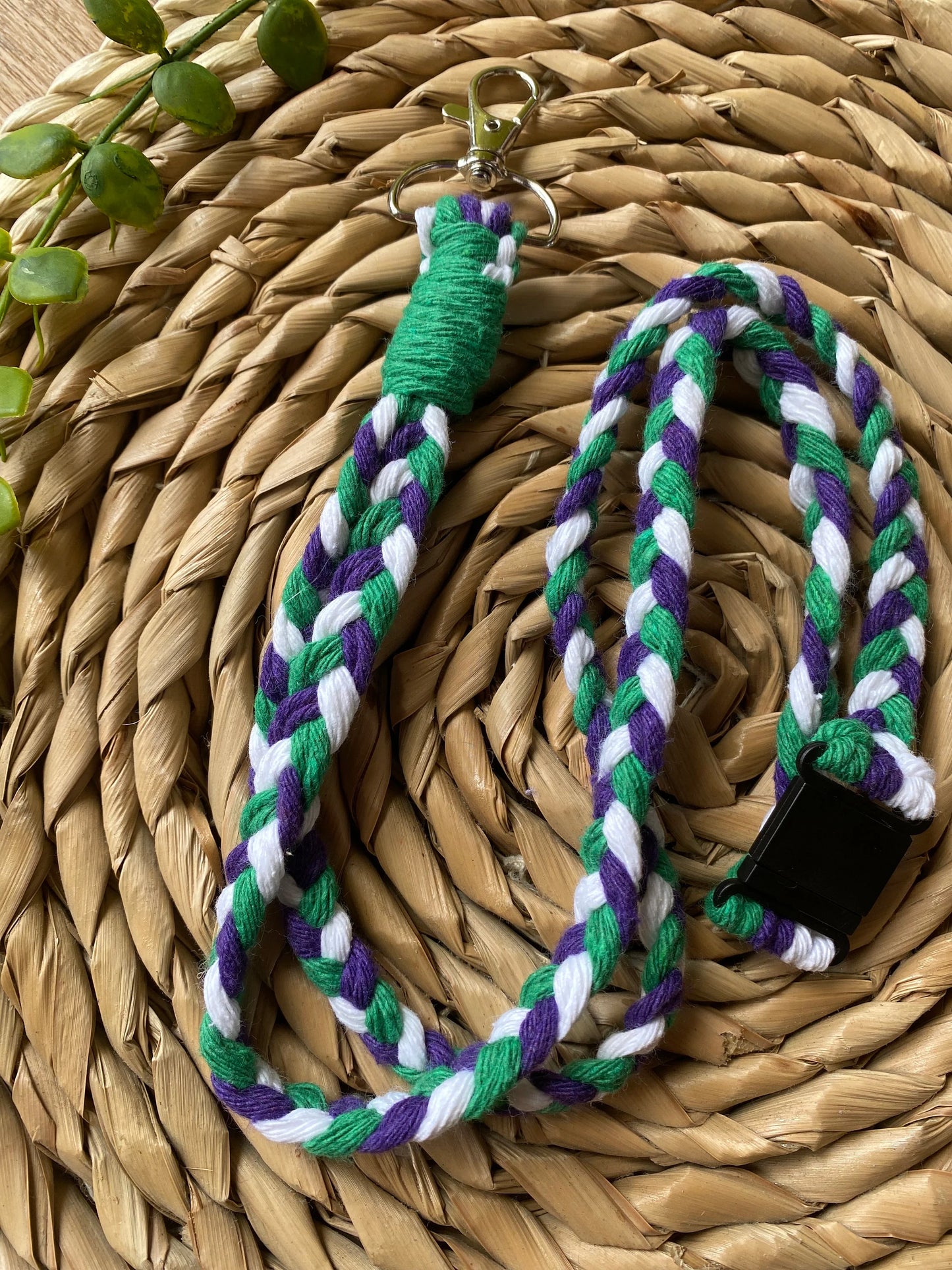 A handmade lanyard with green, purple, and white braided strands lies on a woven circular mat. The Macra-Made-With-Love Eco friendly lanyard badge holder - 34” features a green-wrapped top section with a silver clasp and a black plastic clip at the bottom. Small green plant leaves are visible in the corner.