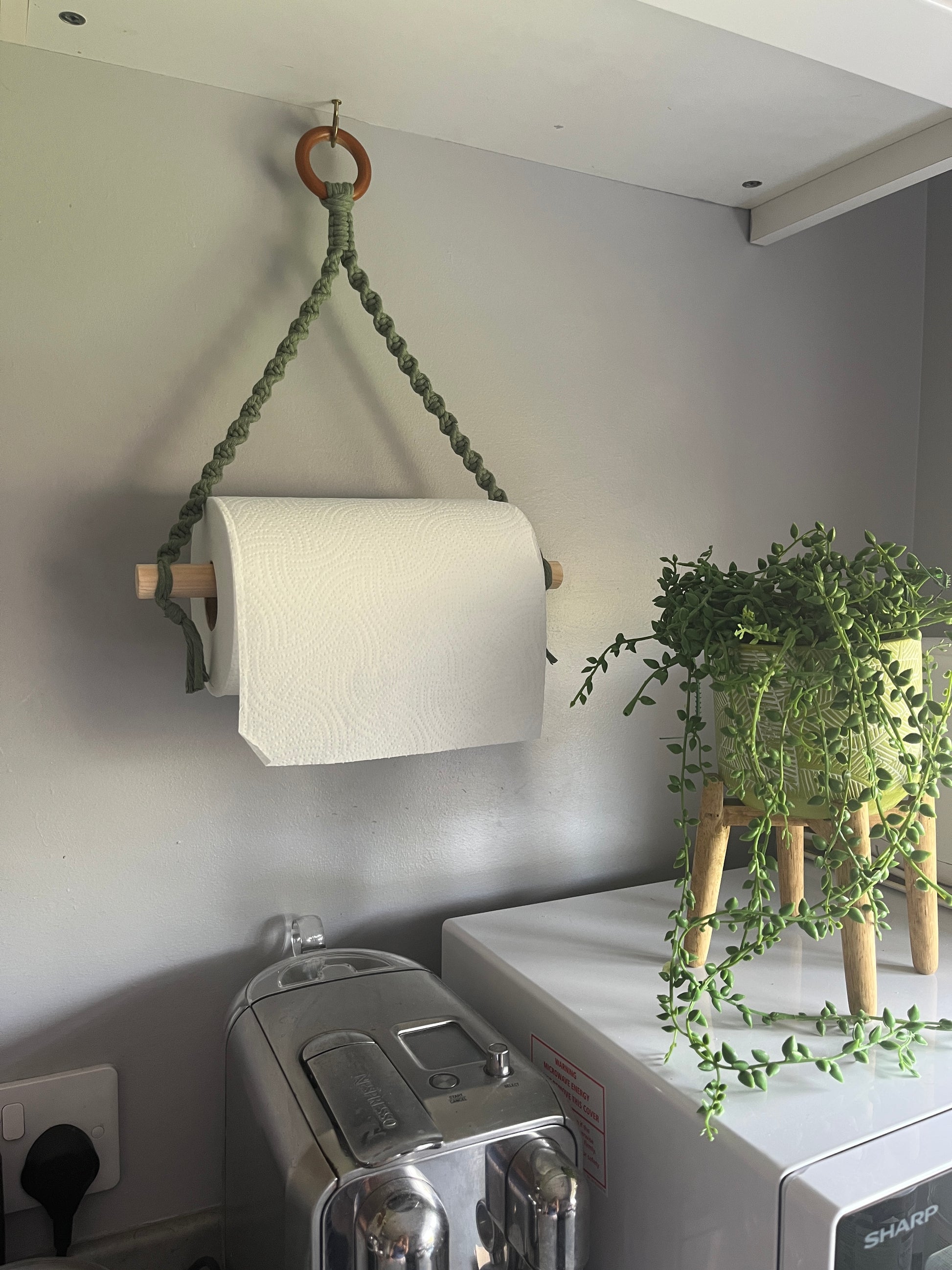 Roll of white paper towel on a macrame wall hanging holder made with sage green yarn and a wooden ring, next to a plant which is placed on top of a white microwave. 