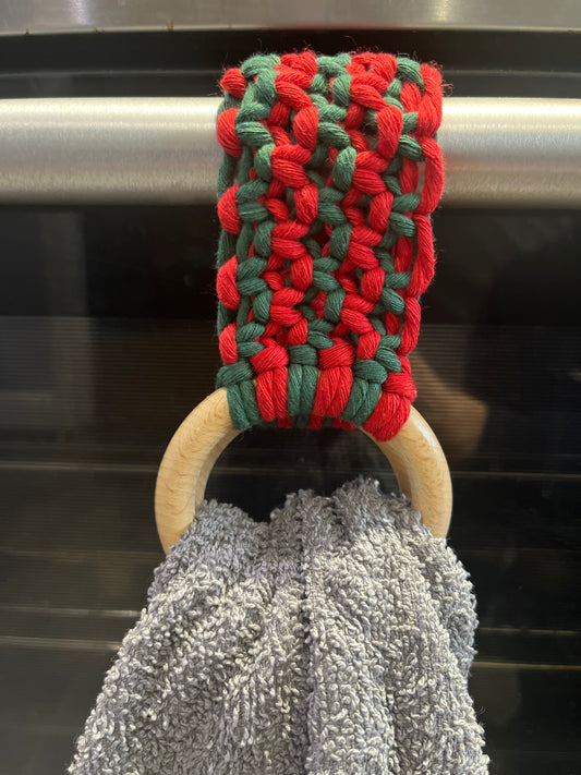 A handmade macrame kitchen tea towel ring holder, made with red and green cotton yarn, and two wooden rings, is looped over a silver oven handle, and holds a grey tea towel between the wooden rings.