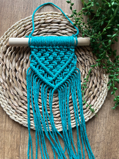 Blue macrame wall hanging on a wooden dowel, laid across a round woven mat with greenery