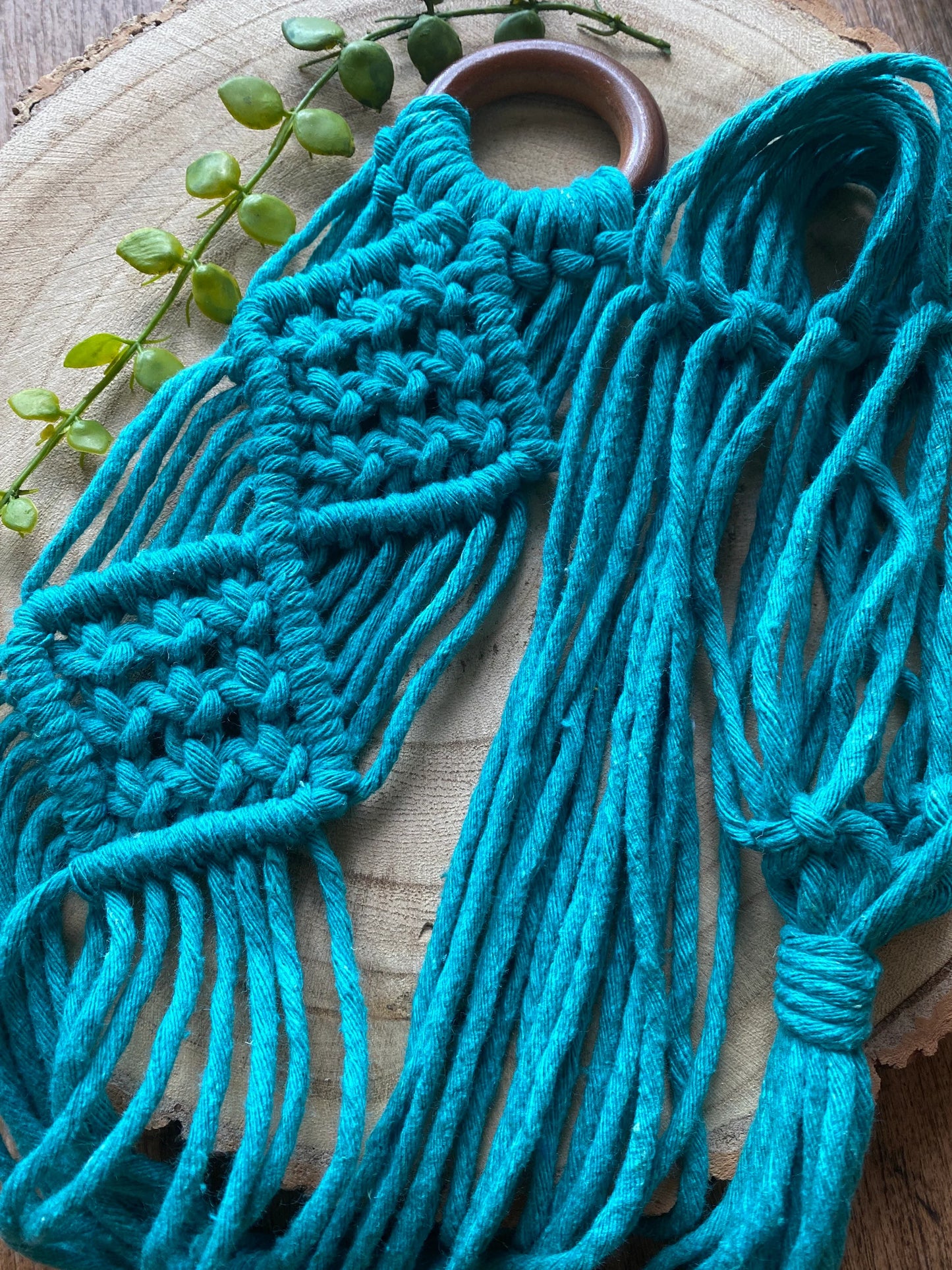 A handmade macrame ukulele wall hanger, made with recycled cotton in turquoise blue, on a repurposed wooden hanging ring. The hanger is laid out on a round wooden surface, and you can see the intricate knot pattern details. 