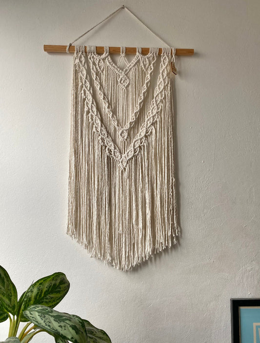 A large statement macrame wall hanging made with natural cotton yarn on a long wooden dowel, with layered knots and boho style. Hanging on a white wall. 