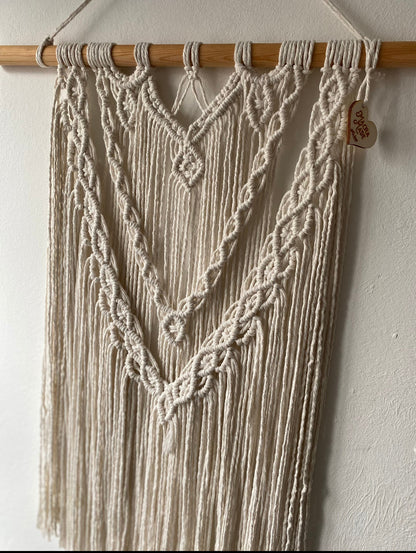 A close up of a large statement macrame wall hanging made with natural cotton yarn on a long wooden dowel, with layered knots and boho style. Hanging on a white wall. 