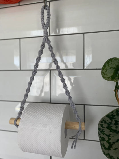 A roll of toilet paper hangs on a wooden dowel suspended by a handmade Macra-Made-With-Love Single toilet roll holder, an eco-friendly decor choice. The background features white subway tiles with grout lines creating a grid pattern. To the right, there is a partial view of a green plant with patterned leaves.