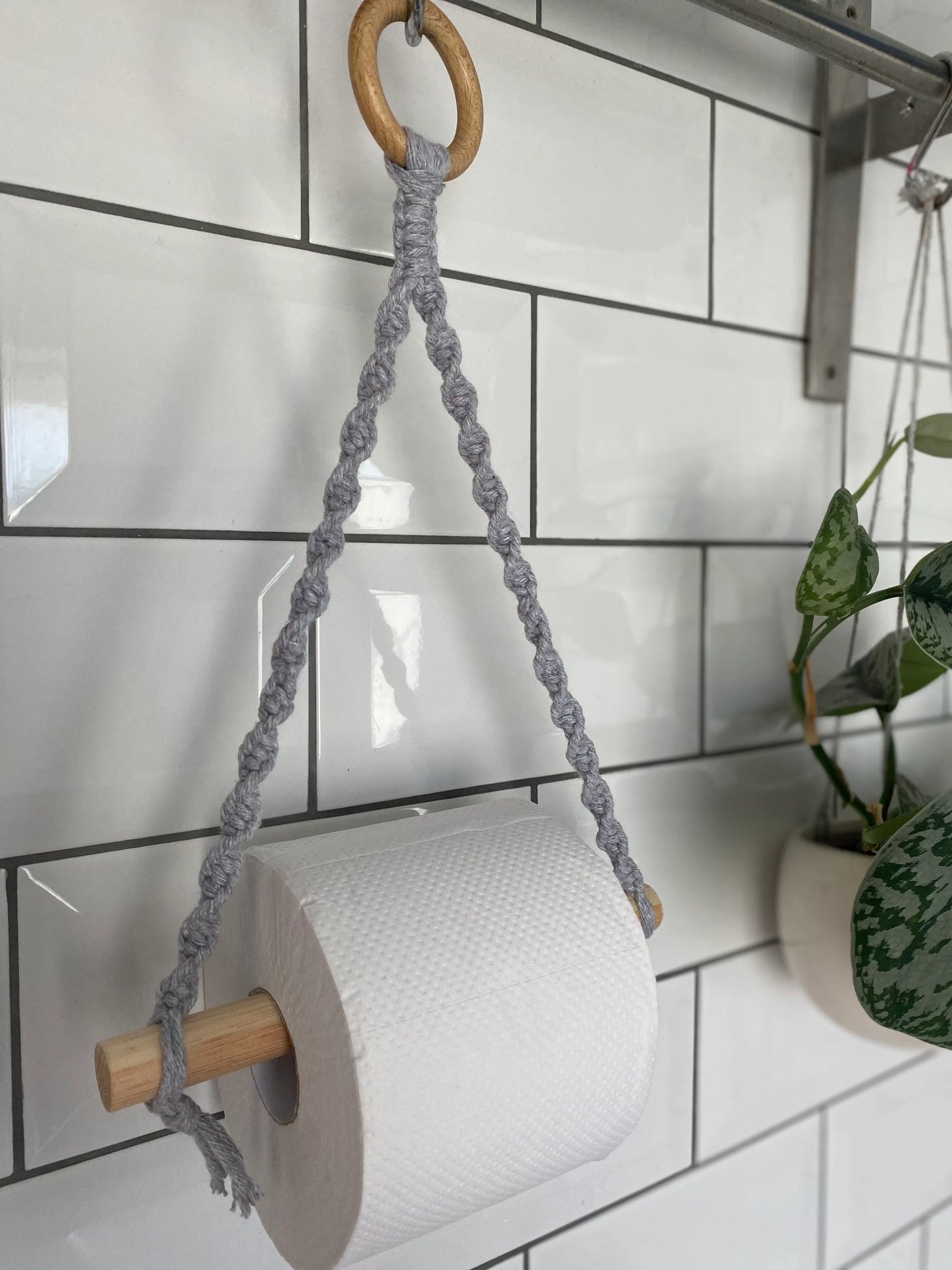 A handmade Macra-Made-With-Love Single toilet roll holder hangs on a wooden dowel, suspended by braided gray rope attached to a wooden ring. This eco-friendly decor is mounted against a white tiled wall, with a potted plant partially to the right.