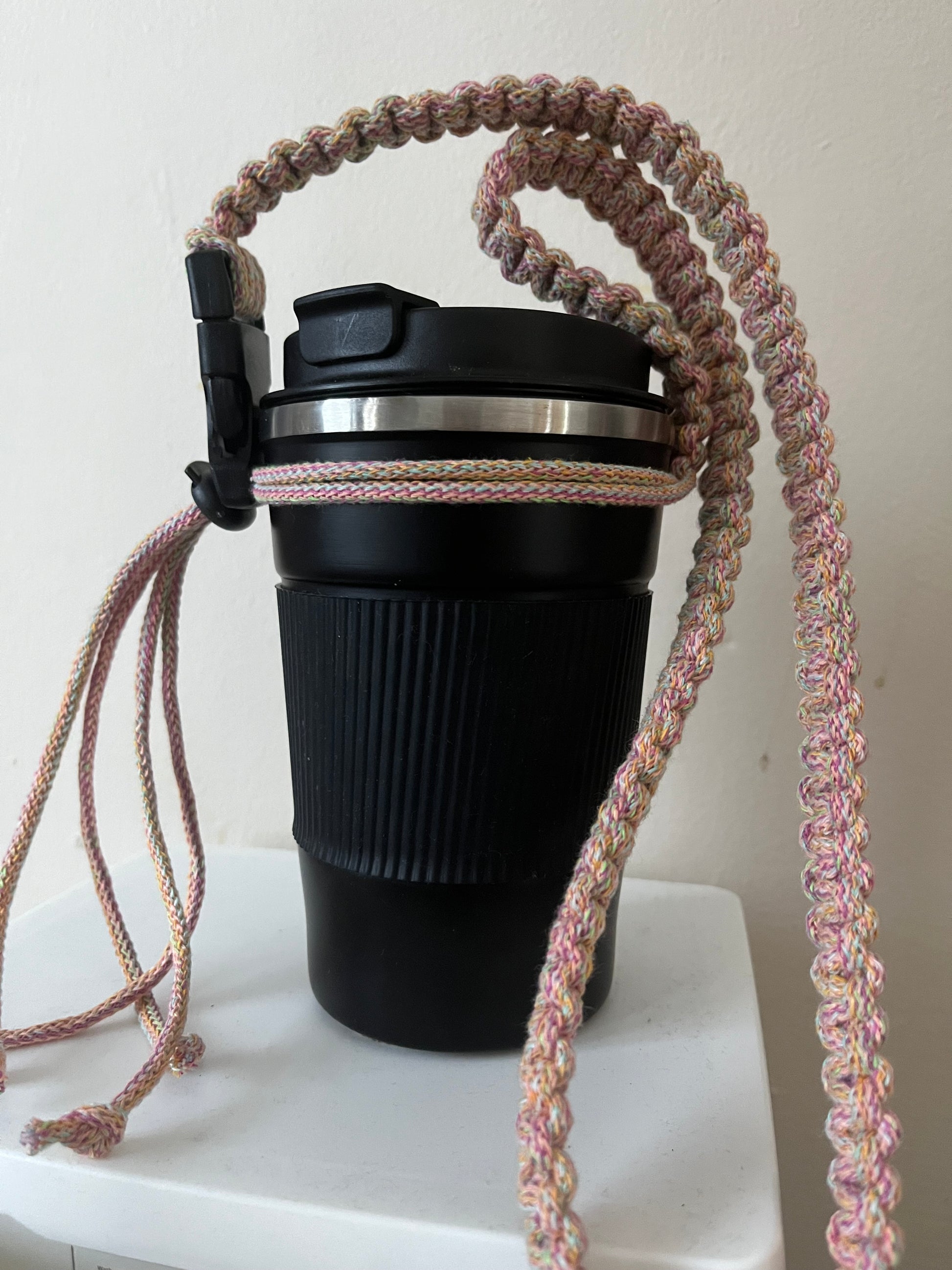 A black reusable coffee cup has a long adjustable coffee cup holder strap made with eco friendly rainbow cotton attached to it. 