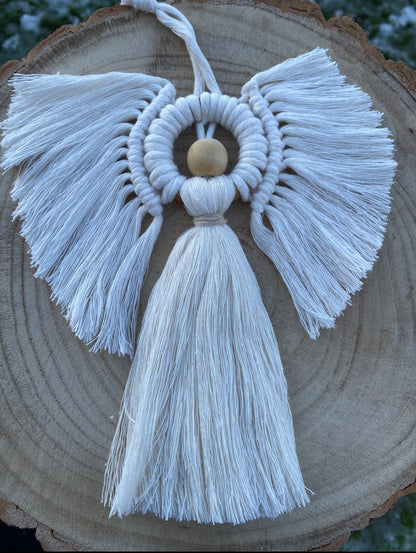 A handmade macrame angel decoration, made with natural yarn and a wooden bead displayed on a round wooden log slice. 