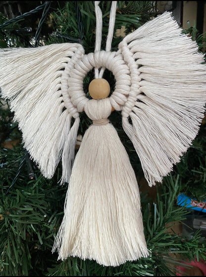 A handmade macrame angel decoration, hanging against the dark green leaves of an artificial christmas tree. 