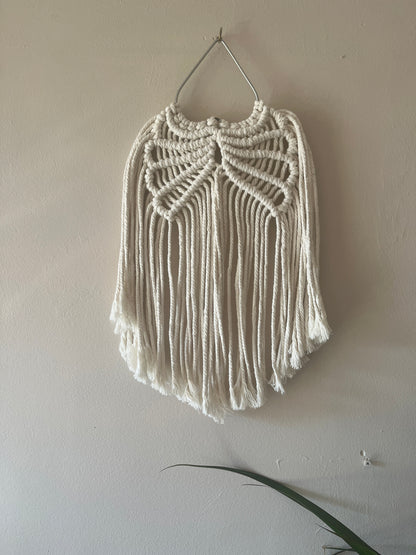 Macrame wall hanging in the shape of wings, made with natural cotton on a metal heat shaped frame, hanging against a plain background. 