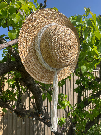 A wicker hat hangs from the branch of a pear tree, with a handmade boho style macrame hat band wrapped around the crown of the hat, with a long tassel. 