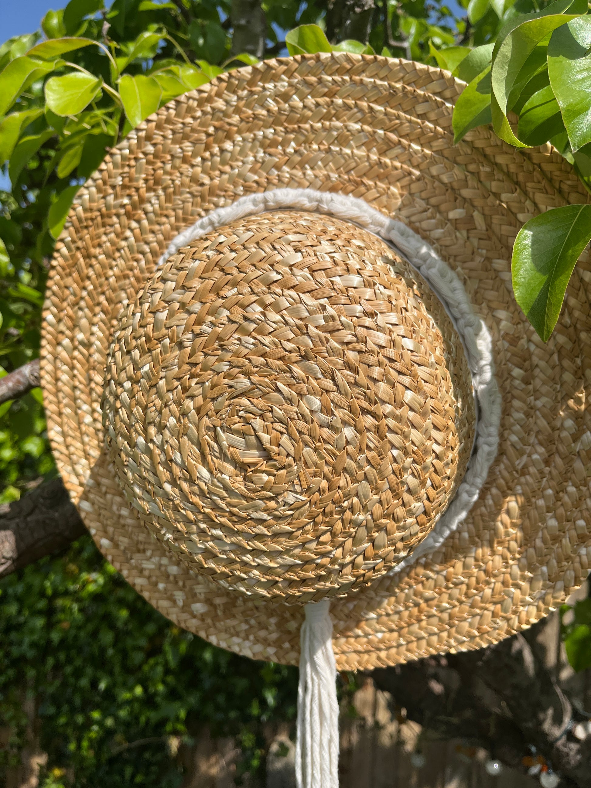 A wicker hat hangs from the branch of a pear tree, with a handmade boho style macrame hat band wrapped around the crown of the hat, with a long tassel. 