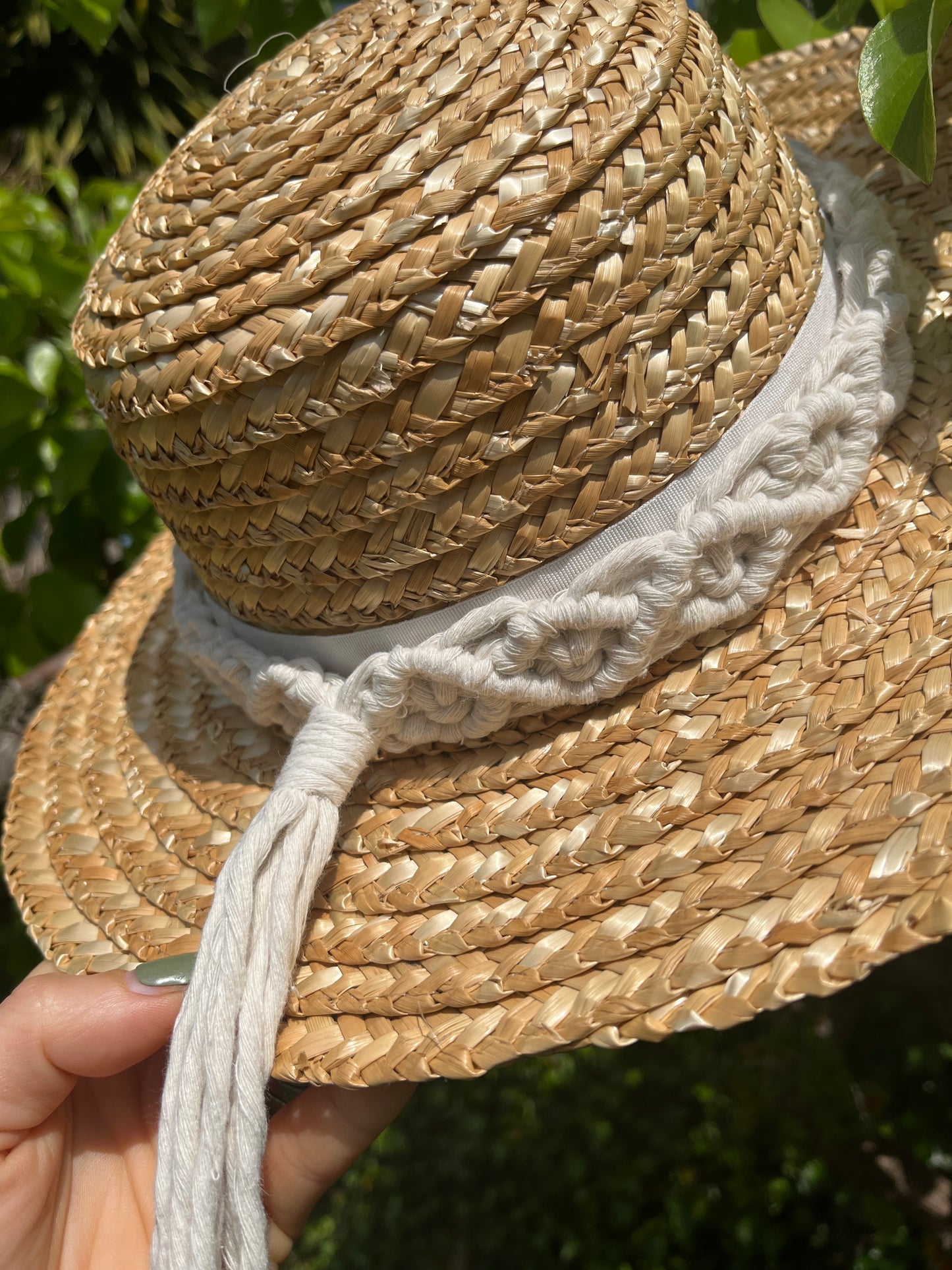 A hand is holding a wicker hat outside, with a tree in the background, there is a handmade boho style macrame hat band wrapped around the crown of the hat, with a long tassel. 
