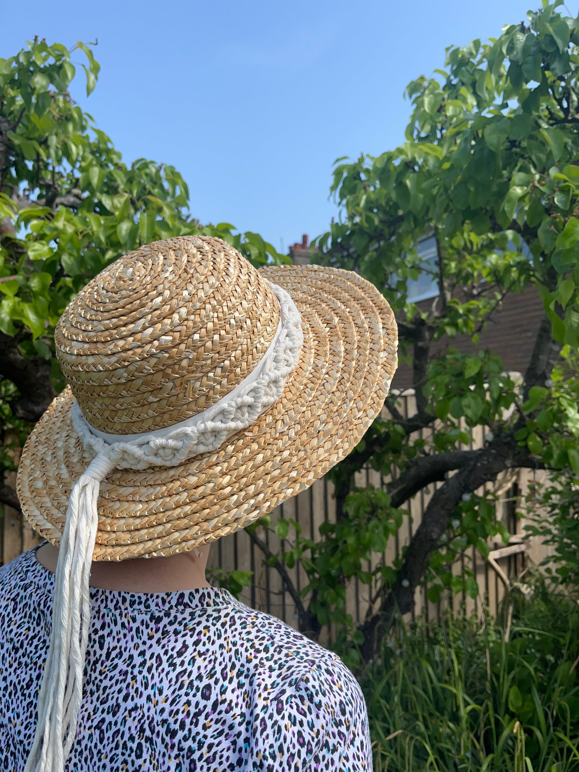 A woman stands outside in front of a pear tree, with her back to the camera. She is wearing a wicker hat with a handmade boho style macrame hat band wrapped around the crown of the hat, with a long tassel. 