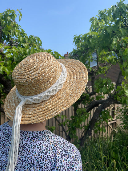 A woman stands outside in front of a pear tree, with her back to the camera. She is wearing a wicker hat with a handmade boho style macrame hat band wrapped around the crown of the hat, with a long tassel. 