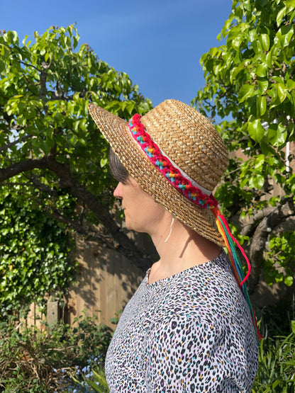 A woman stands outside, facing sideways to the camera, wearing a wide rimmed wicker hat, with a handmade boho style macrame hat band with a long tassel around the crown of the hat. The hat band is made with an intricate knot pattern using rainbow colours of recycled cotton. 