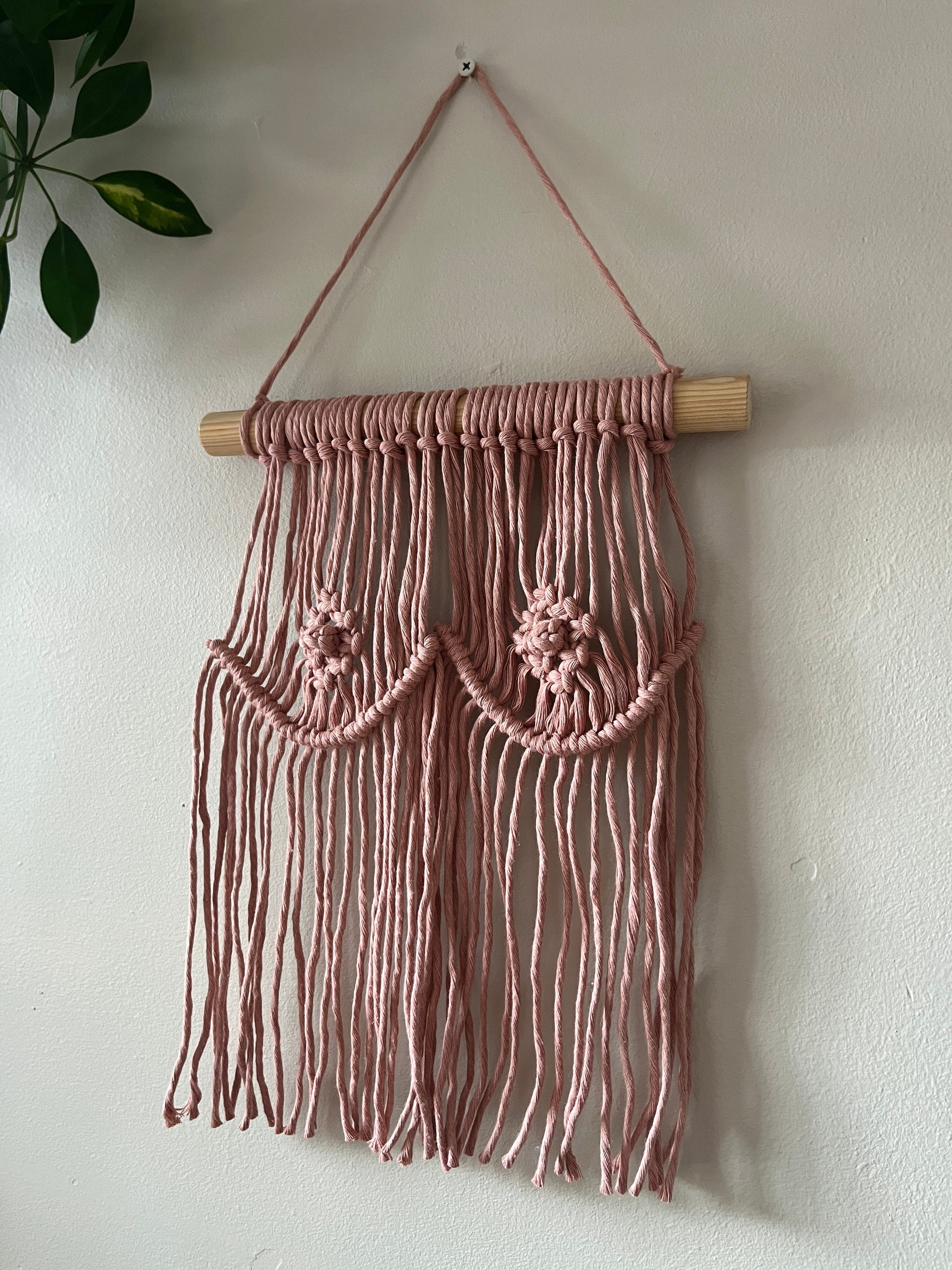 A macrame wall hanging representing boobs, made with blush pink yarn on a wooden dowel, hanging on a beige wall. 