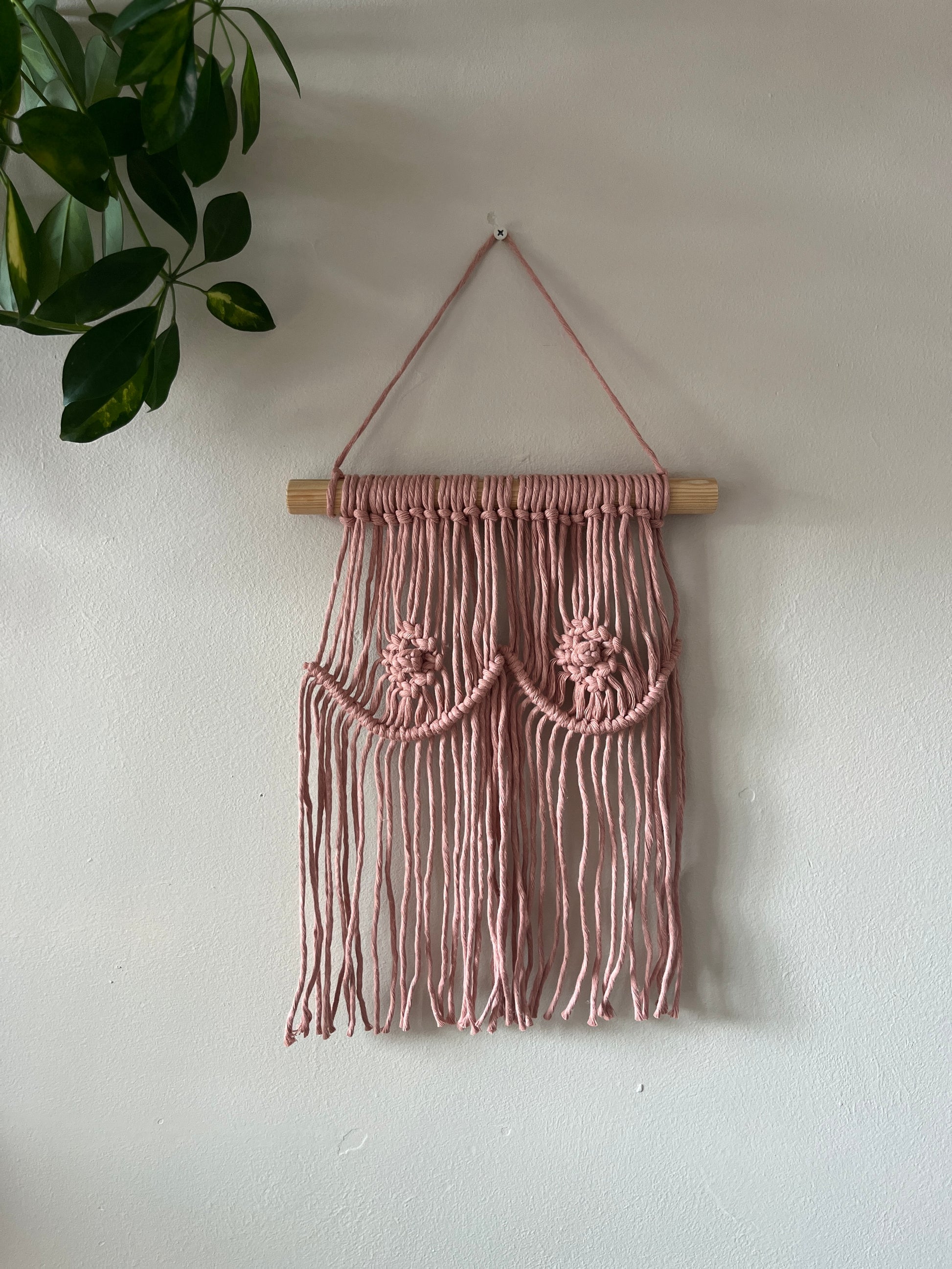 A macrame wall hanging representing boobs, made with pink  yarn on a wooden dowel, hanging on a beige wall. 
