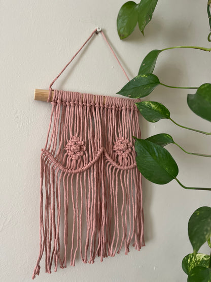 A macrame wall hanging representing boobs, made with blush pink yarn on a wooden dowel, hanging on a beige wall next to a long trailing plant. 