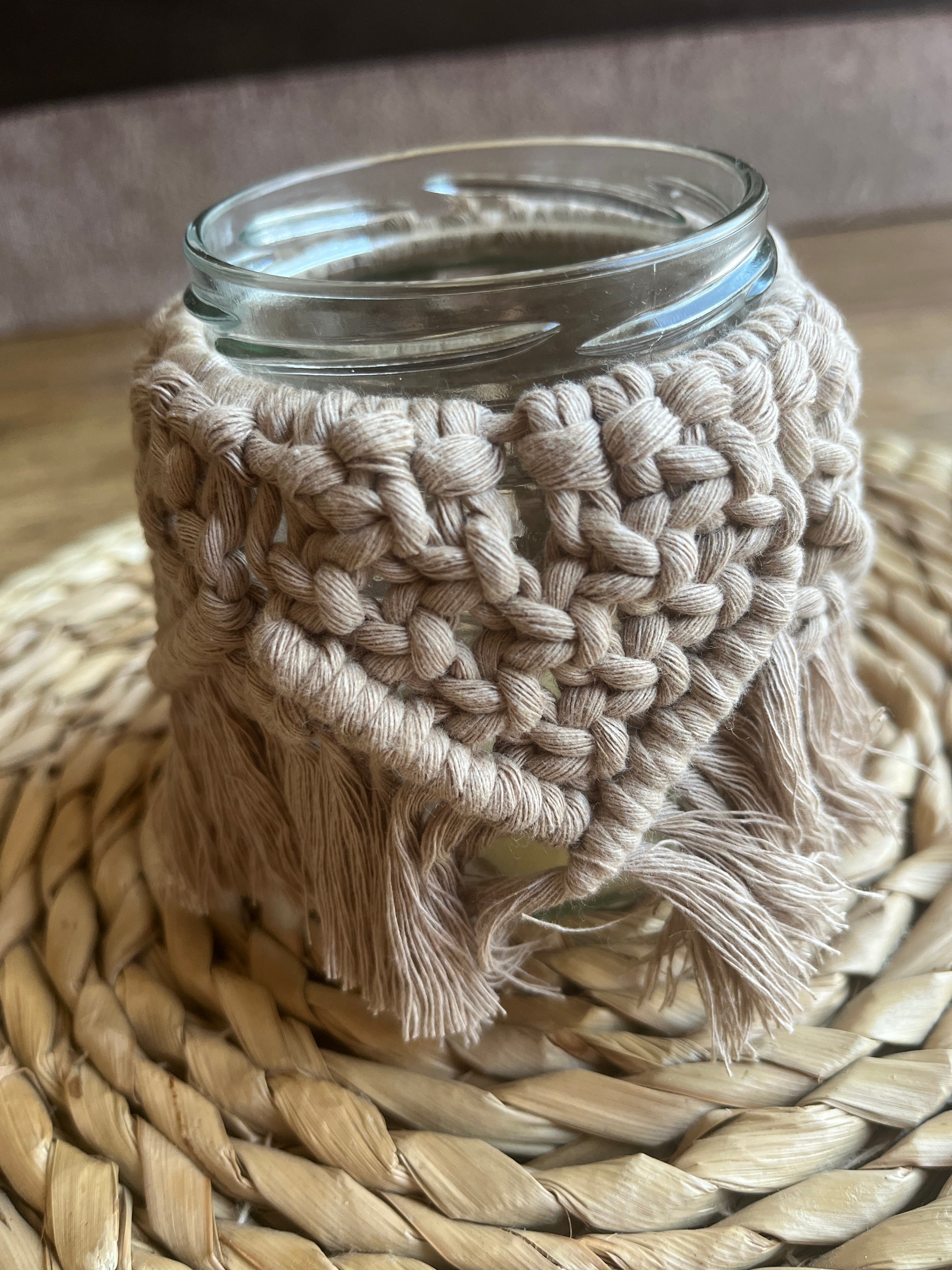 Close up of a glass jar wrapped with cotton yarn with a decorative knotted pattern placed on a wicker surface. 