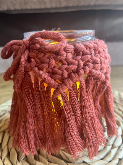 Close up of a glass jar wrapped with terracotta cotton yarn with a decorative knotted pattern and finished with a bow. There is a soft glow from a candle coming from inside the jar. 