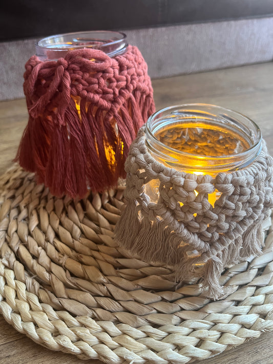 Two glass jars wrapped with cotton yarn with decorative knotted patterns sitting next to each other on a wicker mat. There is a soft glow from a candle coming from inside the jars. 