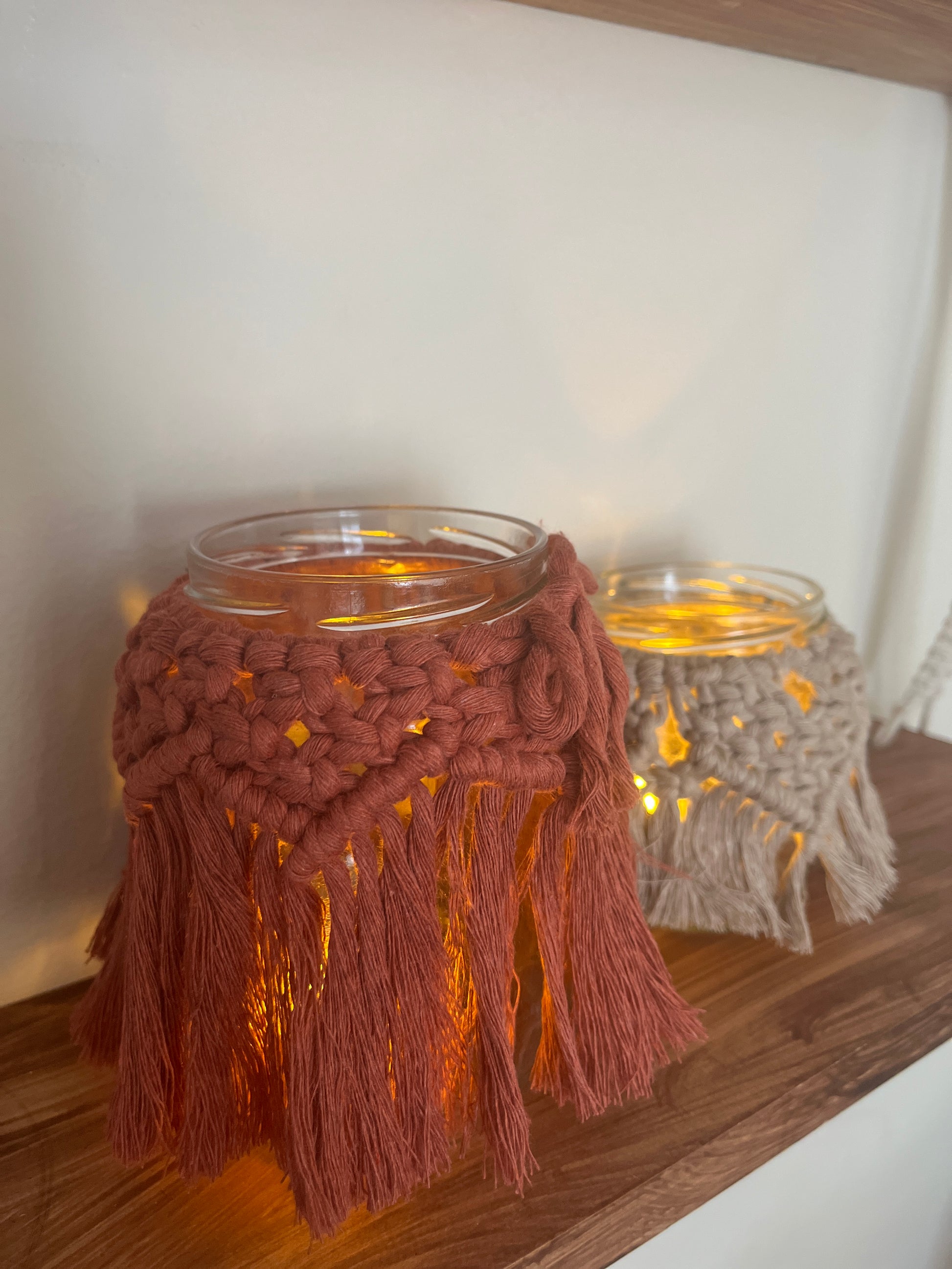 Decorative glass jars with macrame tassels and LED tealights on a wooden shelf.