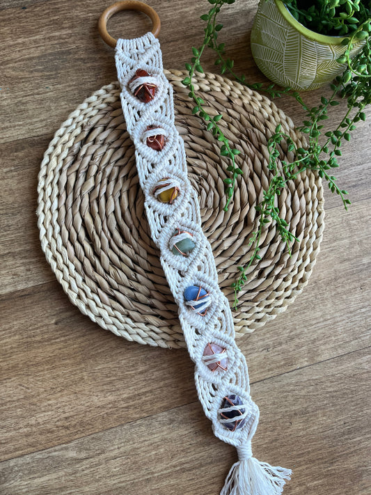 Long macrame wall hanging, featuring 7 gemstones to represent the chakras, held together with copper wire, and wrapped in natural recycled cotton yarn using a diamond knot pattern. finished with a repurposed wooden hanging ring. Displayed on a round wicker mat next to long green plant vines. 