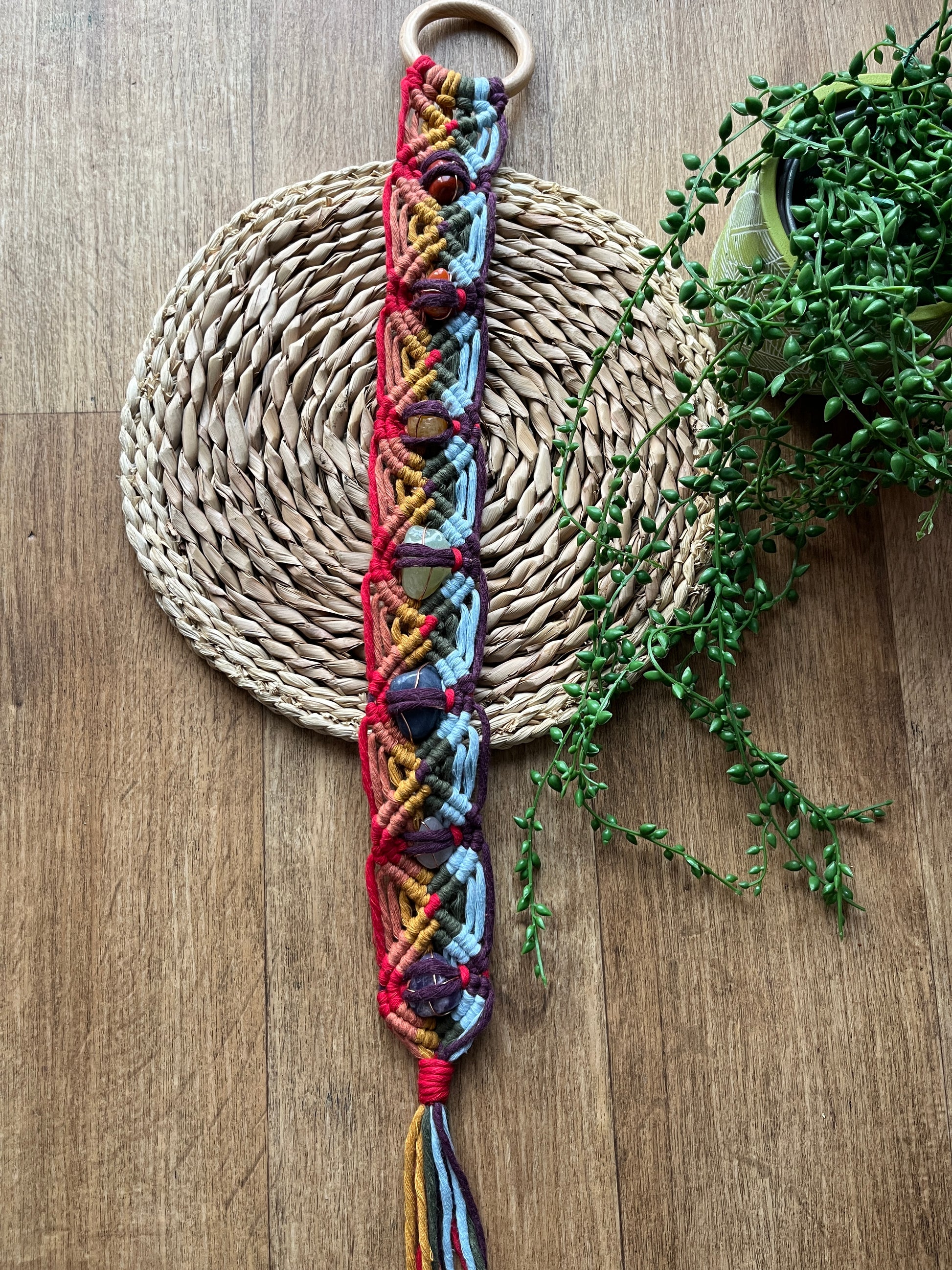 Colourful macrame chakra gemstone wall hanging on a woven mat with greenery