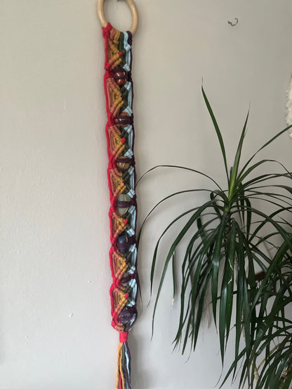 Colourful macrame chakra gemstone wall hanging on a beige wall with a plant in the corner 