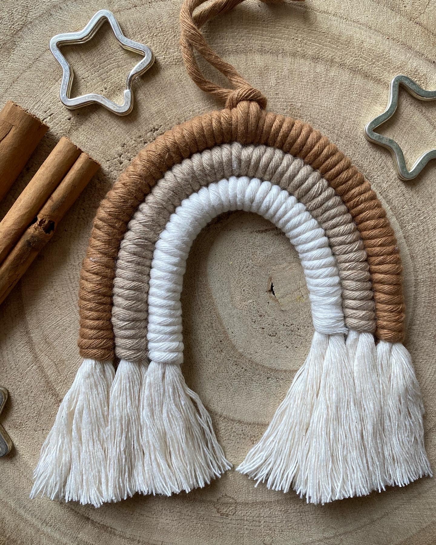 A handmade macrame rainbow Christmas decoration in brown, beige and white yarn with white tassels, displayed on a wooden surface with beads and cinnamon sticks for decoration. 