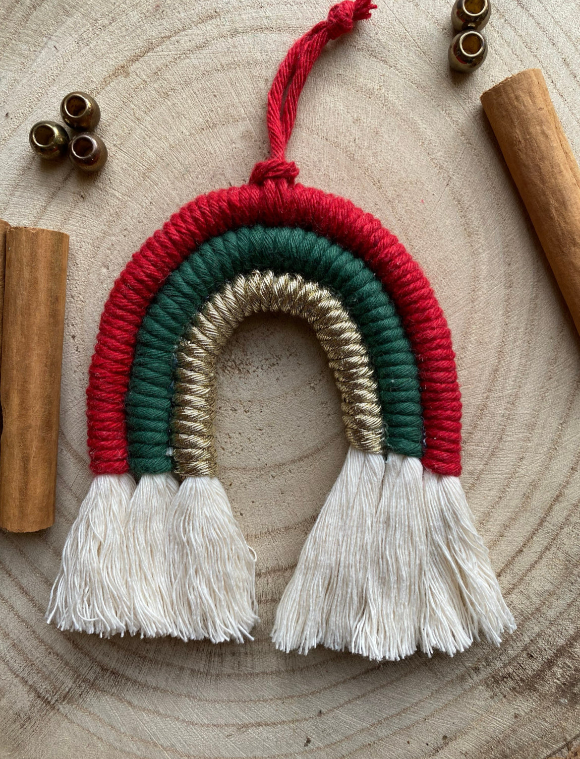 A handmade macrame rainbow in festive christmas colours of red, green and gold,  on a round wooden surface surrounded by small beads and cinnamon sticks. 