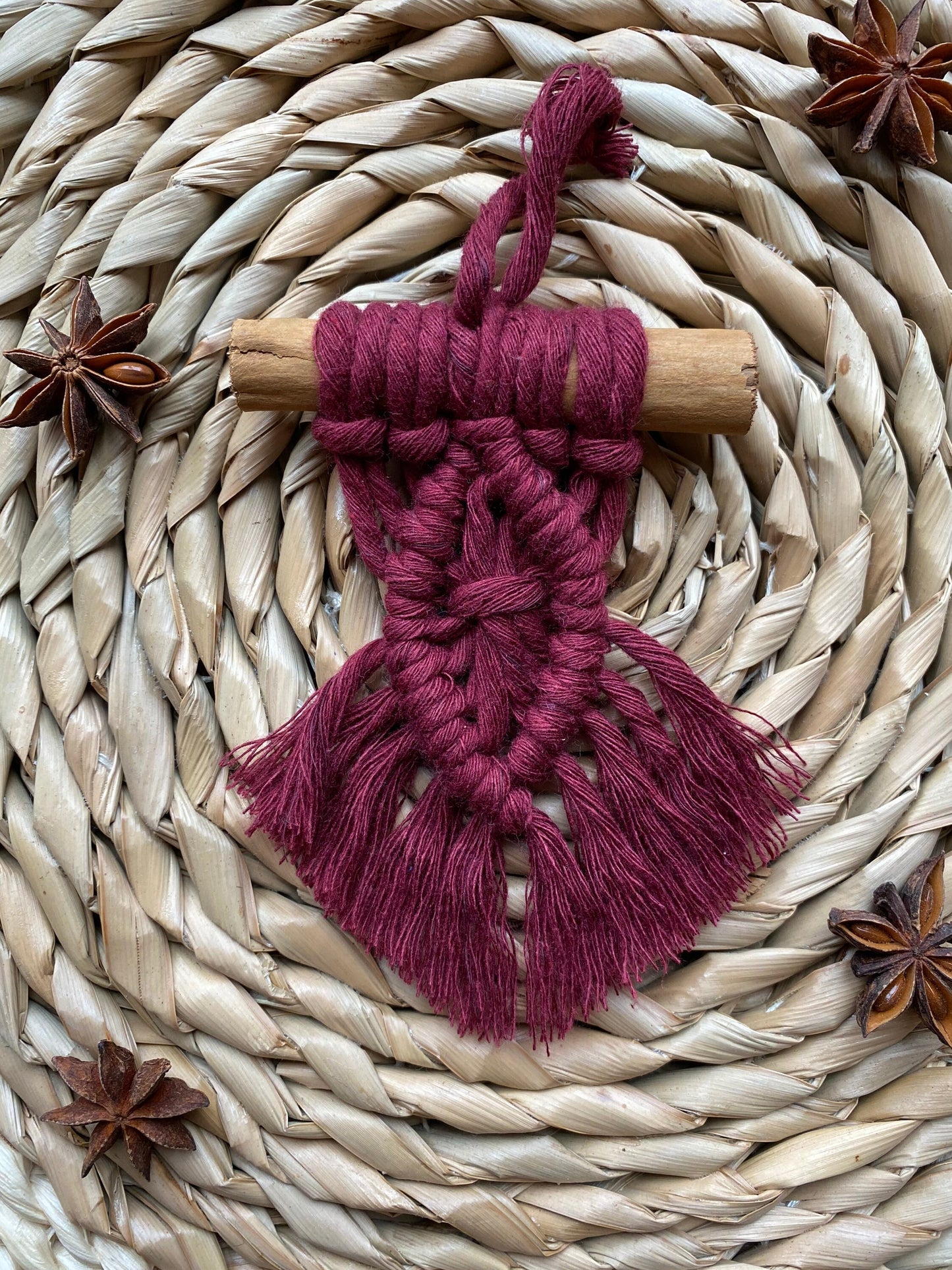 A handmade macrame christmas tree decoration made with deep red recycled cotton and a real cinnamon stick, laid out on a round wicker mat surrounded by star anise for decoration.