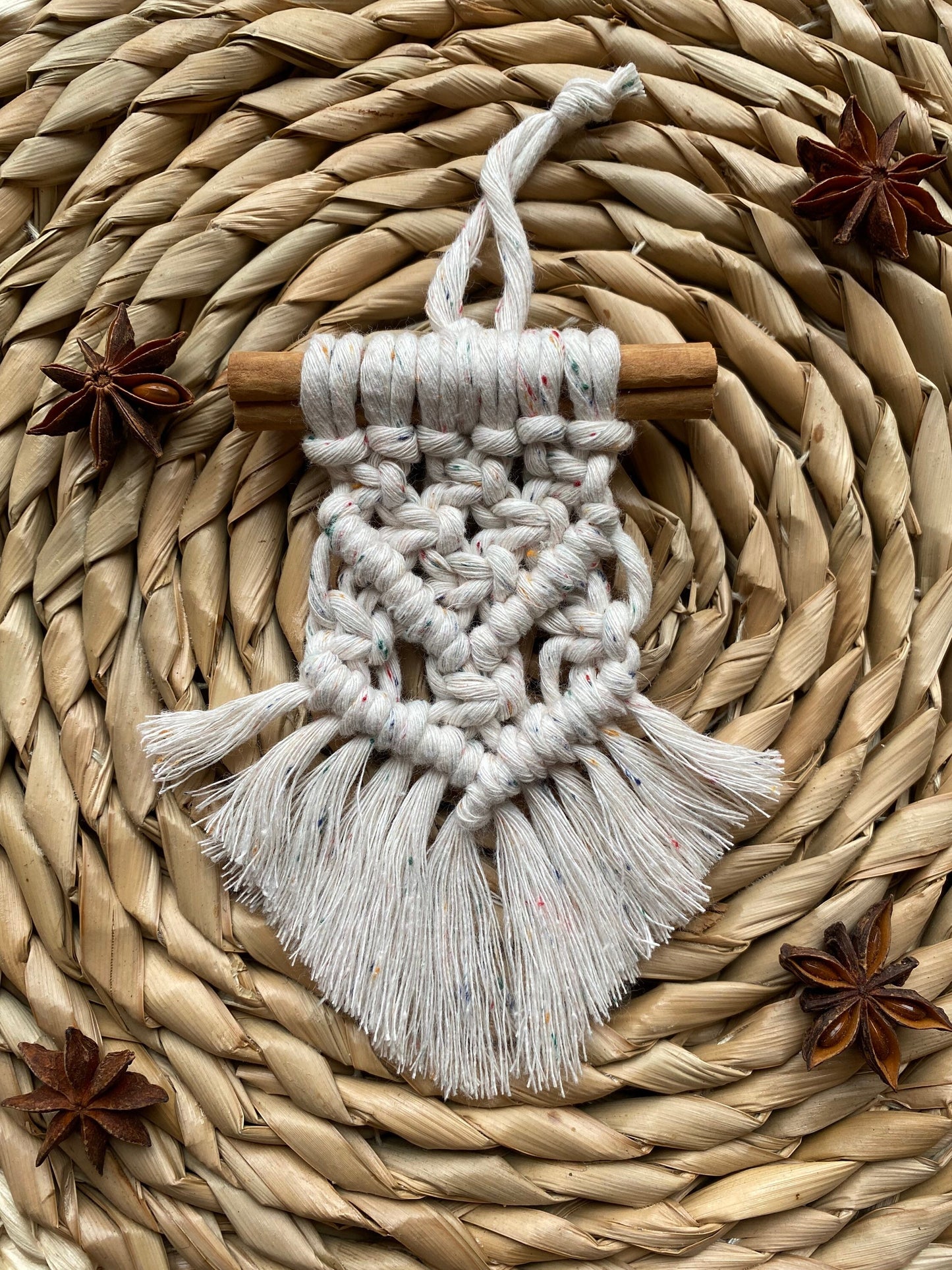 A handmade macrame christmas tree decoration made with recycled cotton and a real cinnamon stick, laid out on a round wicker mat surrounded by star anise for decoration.