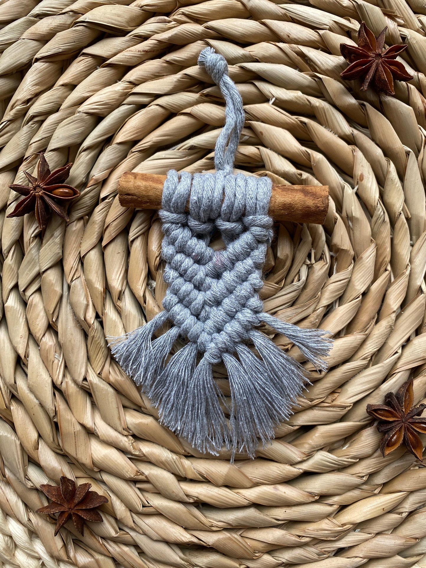A handmade macrame christmas tree decoration made with recycled cotton and a real cinnamon stick, laid out on a round wicker mat surrounded by star anise for decoration.