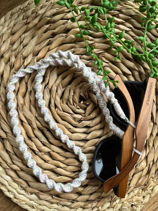 A pair of dark sunglasses with wooden arms are placed on a wicker surface with a handmade macrame eyewear holder chain, made with recycled cotton yarn, attached to them. 