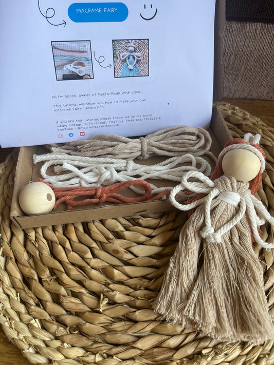 A cardboard box filled with yarn and a wooden bead, sits on a wicker mat with a macrame fairy decoration positioned at the front. 