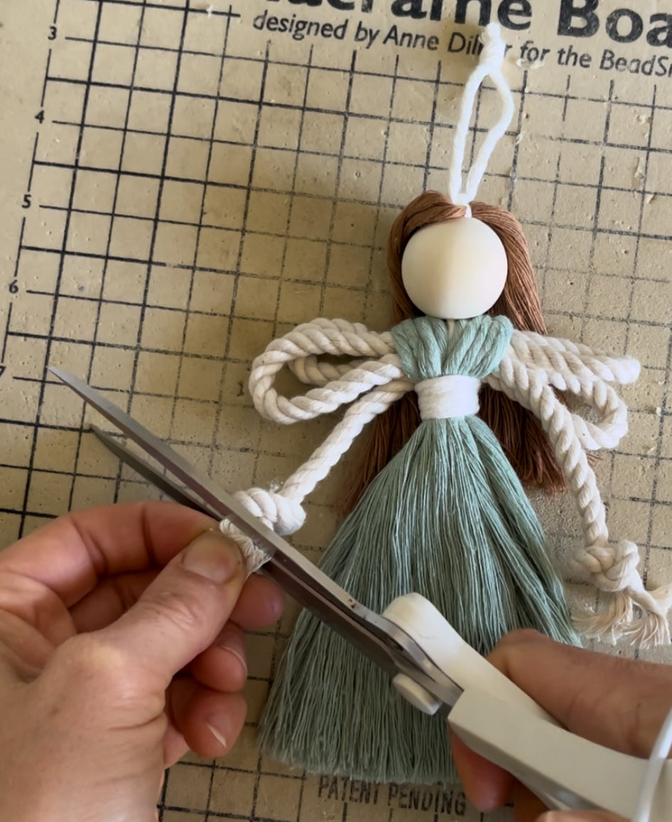 Hands working on a macrame fairy craft kit, cutting rope with large white scissors on a grid background. 