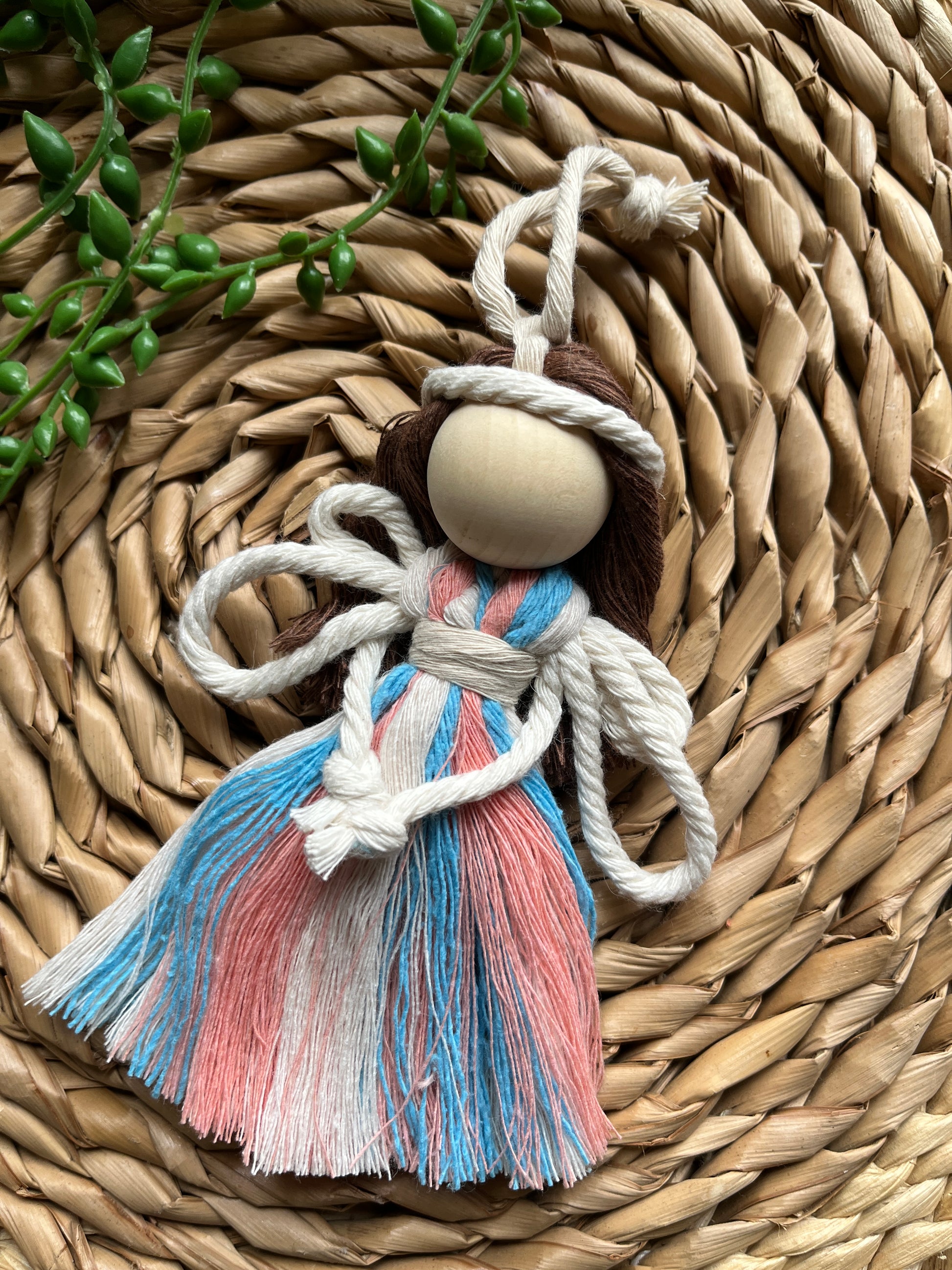 A handmade macrame fairy ornament, with recycled cotton dress in blue pink and white, white rope wings and a large wooden bead, laid on a round wicker mat with some green plant vines.