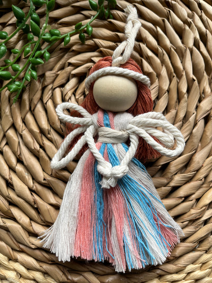 A handmade macrame fairy ornament, with recycled cotton dress in blue pink and white, white rope wings and a large wooden bead, laid on a round wicker mat.