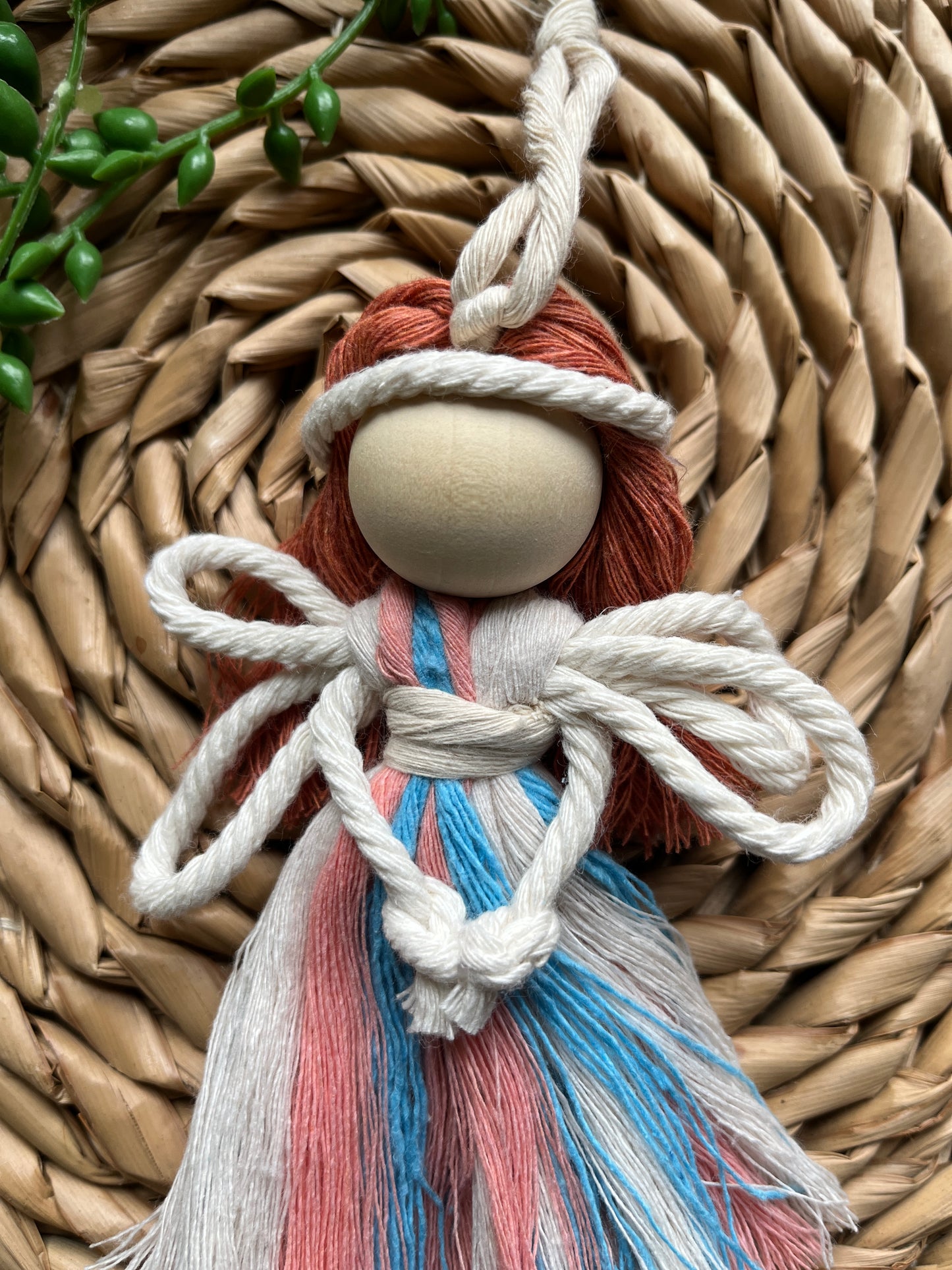 Close up of a handmade macrame fairy ornament, with recycled cotton dress in blue pink and white, white rope wings, a large wooden bead and red cotton hair, laid on a round wicker mat with some green plant vines.