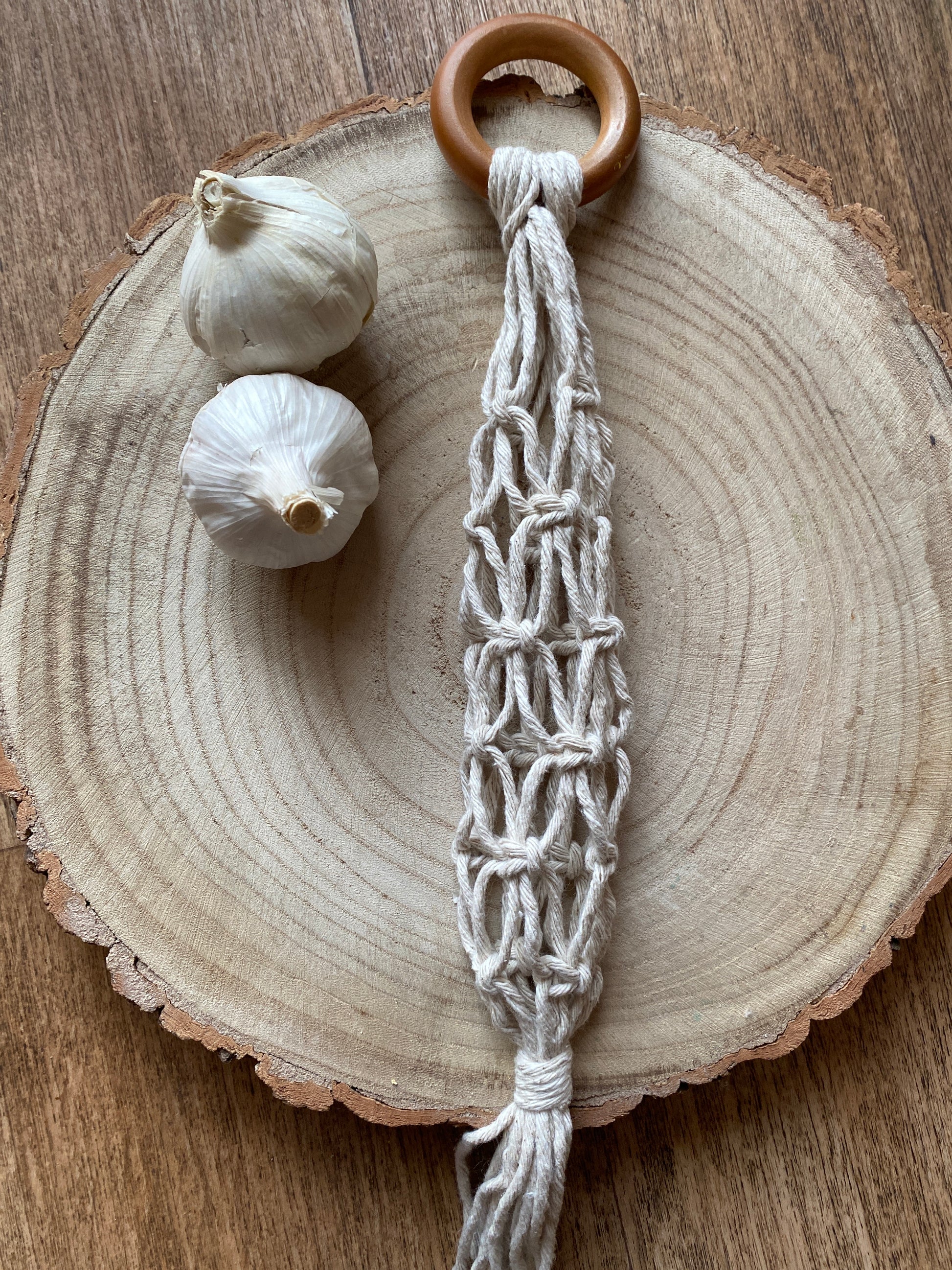 A handmade macrame garlic hanger made with natural cotton yarn and a wooden ring, laid on a round wooden board next to two fresh garlic bulbs. 