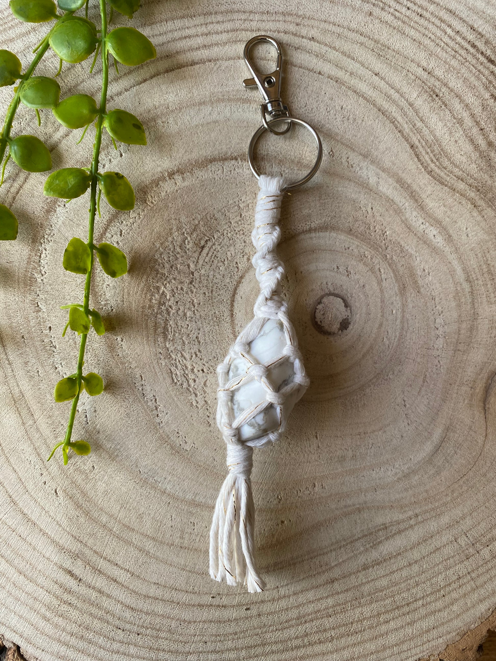 A handmade gemstone Keyring featuring a white howlite stone encased in intricate white cord knots. The keyring has a metal lobster clasp and is placed on a wooden surface with a green plant vines in the upper left corner. 