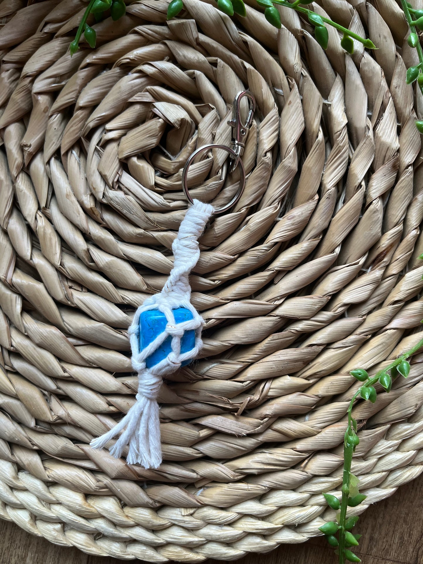 A handmade macrame gemstone keyring, made with natural coloured cotton yarn wrapped around a blue howlite gemstone, and metal split ring clasp, on a wicker surface.