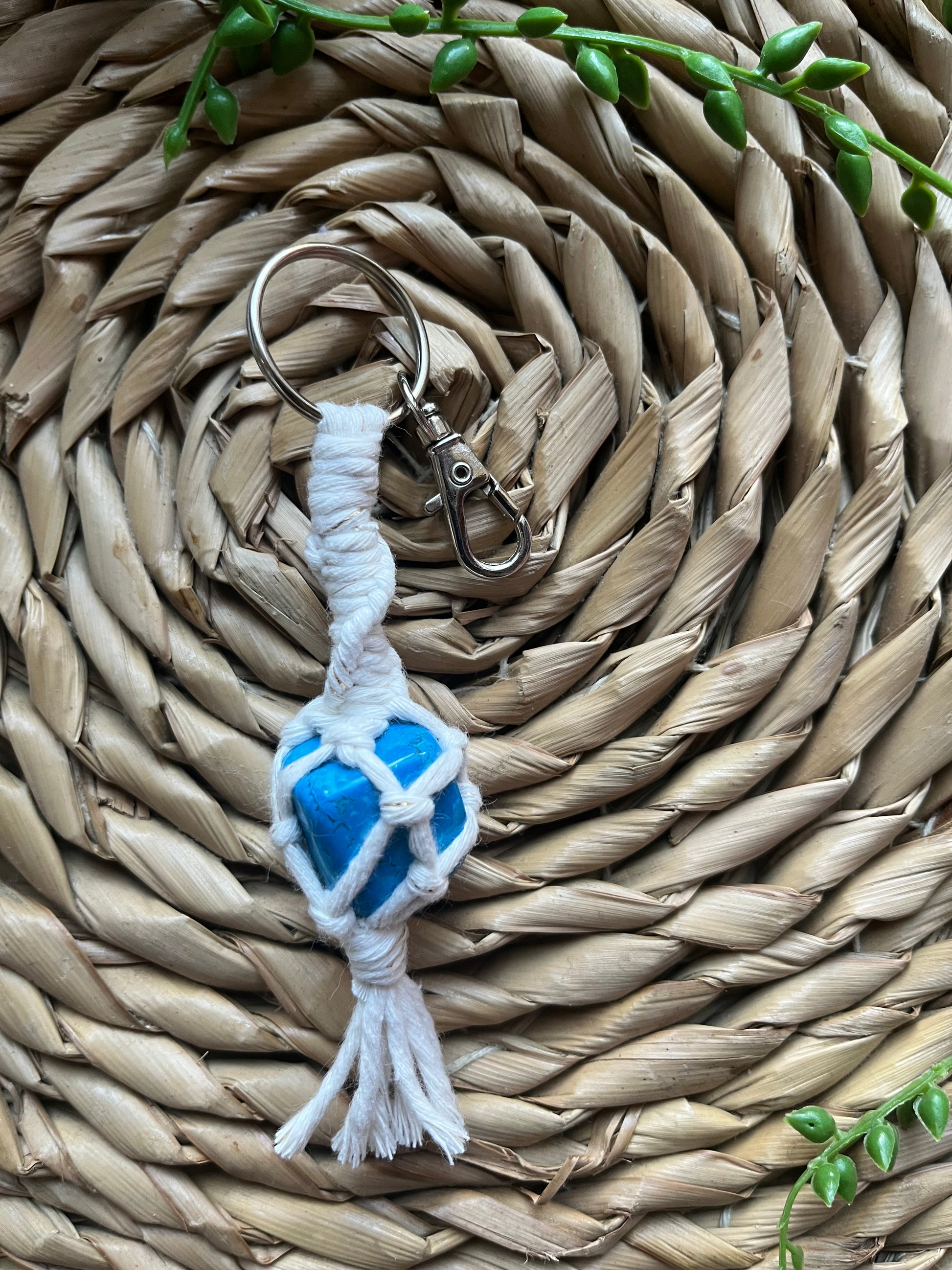 A handmade macrame gemstone keyring, made with natural coloured cotton yarn wrapped around a blue howlite gemstone, and metal split ring clasp, on a wicker surface.