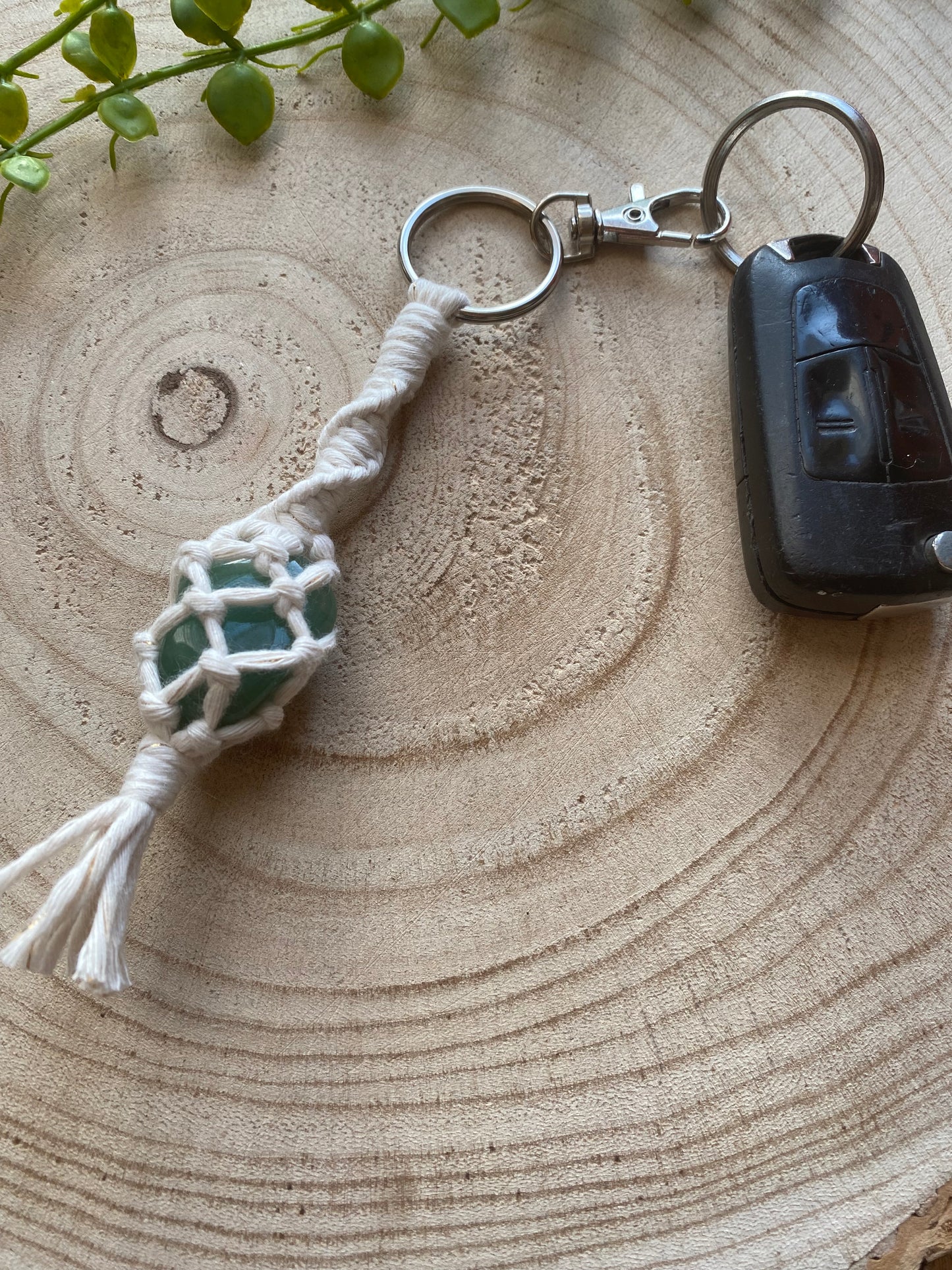 A handmade macrame gemstone keyring, made with natural coloured cotton yarn wrapped around a green quartz gemstone, and metal split ring clasp, on a wooden surface attached to a single car key. 