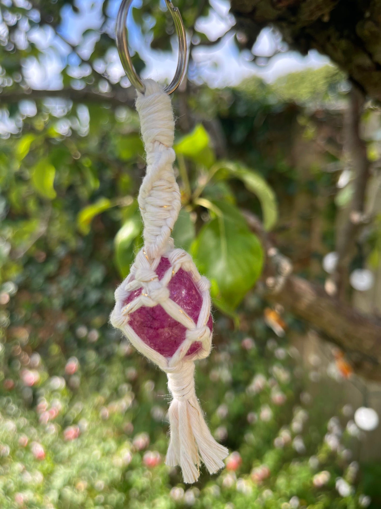 A handmade macrame gemstone keyring, made with natural coloured cotton yarn wrapped around a pink crackle quartz gemstone, displayed hanging outside amongst greenery. 